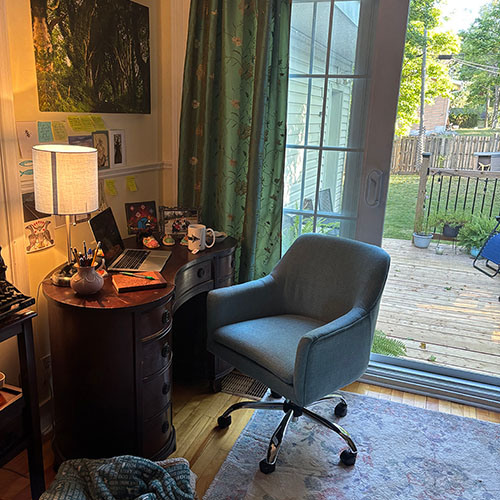 A grey office chair is positioned in front of an antique desk with a laptop and a cup on it. The desk is also adorned with a picture and a lamp. Through a sliding patio door the room opens into a large green yard. Some potted plants can be seen on the edge of the deck by the railing.