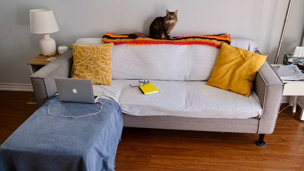 A grey sofa with a white blanket and yellow throw cushions on it. A laptop resting on an ottoman sits in front of the sofa inside Sean Minogue's writing studio.