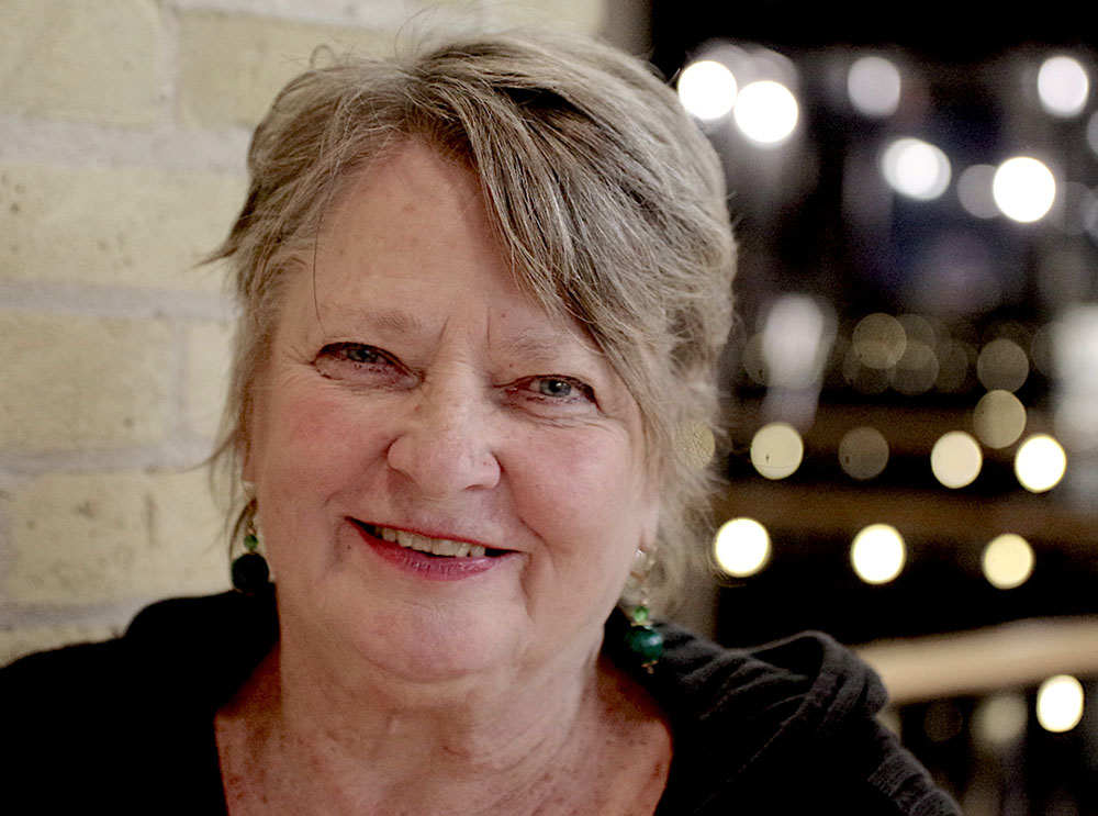 Di Brandt posing in front of a brick wall with lights in the background.